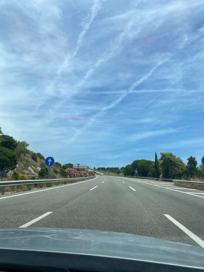 The image displays a forward-facing view from a vehicle on a multi-lane highway in Sant Pol de Mar, Spain, under a clear blue sky. The sky features scattered white clouds and multiple distinct white contrails, some intersecting, with a faint pinkish hue visible within clouds near the left-center horizon. The asphalt highway surface is dry, marked with white dashed and solid lines defining at least two lanes in the direction of travel. Silver-colored metal guardrails line both sides of the road.

On the left side, an ascending, vegetated embankment is visible, covered with green trees, shrubs, and prominent clusters of pink flowering plants. A circular blue road sign with a white upward arrow, indicating a mandatory straight direction, is mounted on this embankment. On the right side, a similar embankment is covered with dense green foliage, including tall, slender trees and broader-leafed trees. A partial view of another identical blue arrow sign is visible on the far right edge. In the far distance on the right lanes, two light-colored vehicles are discernible. The top edge of a dark grey dashboard or hood of the observing vehicle is visible across the bottom of the frame. No people are visible.