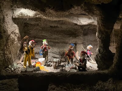The image depicts a dark, cave-like setting, possibly a mine, with rough, textured walls and formations of what appears to be crystal or salt deposits illuminated by artificial light. The scene is populated by several mannequins dressed as miners. These figures are positioned in various stances, engaged in activities suggestive of mining work.

The mannequins are dressed in traditional miner's attire, including plaid shirts, vests, sturdy pants (one pair is bright yellow), and hats. Some are holding tools such as picks, shovels, and a lantern. One figure appears to be using a broom or brush. The mannequins have white beards and are depicted with lifelike facial features. The overall impression is a diorama or exhibit designed to represent mining life.

Notable details include the dramatic lighting that casts shadows and highlights the textures of the cave and the mannequins. The presence of what looks like crystalline formations suggests the environment might be a salt mine or a geode cave. The figures are depicted in a way that conveys a sense of labor and industry. The orientation of the image is upside down, which might be a stylistic choice or an artifact of how the photo was taken.

The setting is identified as Tolk, Germany, which, combined with the visual cues, points to this being a display within a tourist attraction or museum related to mining or local folklore in that region. No visible text is present in the image. The emotions conveyed are primarily related to the stoic nature of hard work, with a touch of the fantastical due to the mannequin figures.