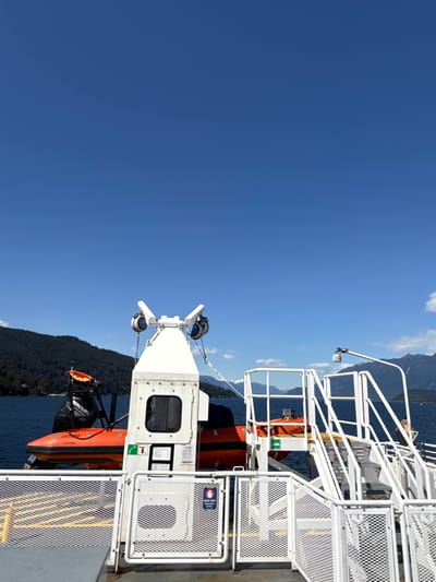 The image depicts a section of a ship or ferry in Bowen Island Municipality, Canada. The sky is clear and blue, with a hint of distant mountains. The ship's structure is white with metal railings and staircases. A lifeboat, painted orange, is visible. There are also mechanical devices on the ship. Water and treed land are in the background.