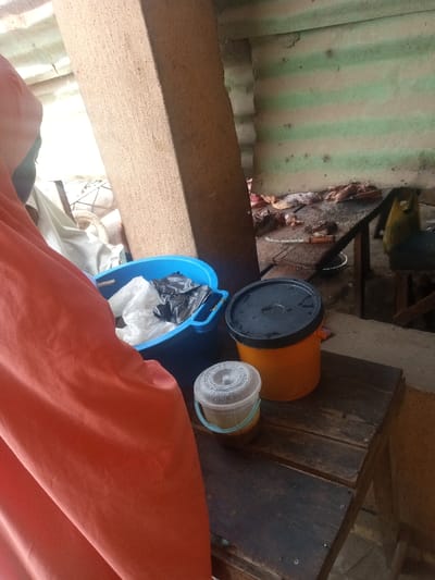 This image captures a close-up scene within an indoor or semi-covered outdoor commercial or domestic space, specifically in Unguwa Uku Kauyen Alu, Nigeria.
In the immediate foreground, to the left, a large segment of an orange fabric object, possibly a garment or drape, occupies the left third of the frame. Adjacent to it and centrally positioned, a prominent, vertical, rectangular pillar constructed of rough-textured light brown material extends from the bottom to the top of the image.
To the left of the pillar, a bright blue plastic basin contains an assortment of white and black plastic bags. To the right of the pillar, a dark wooden surface, possibly a low table or stool, supports two containers. The larger container is an orange bucket with a black lid. Next to it is a smaller, translucent plastic container with a clear, ridged lid, holding a yellowish-brown substance.
In the background, behind the pillar and containers, a wall made of corrugated metal sheeting is visible, characterized by alternating horizontal stripes of green and a faded lighter color. Beneath this wall, a dark wooden table is laden with numerous pieces of raw meat and bones. A knife with a light-colored handle is partially visible on this table amidst the meat. Further back and to the right, indistinct objects, including a yellow bag, are present. The scene indicates a food preparation or market area, likely a butchery. No individuals are explicitly visible or engaged in direct action within the frame.