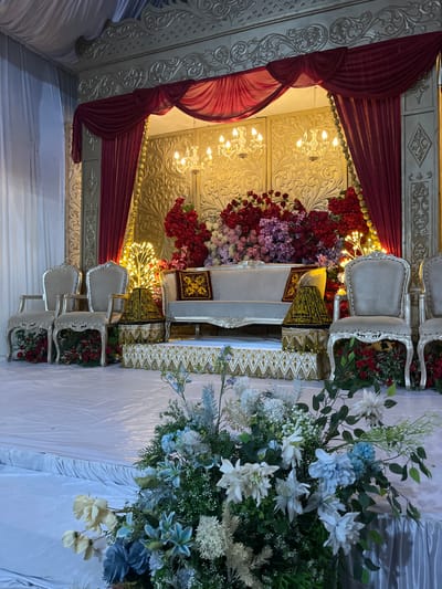 This is a detailed, forensic, matter-of-fact description of an indoor wedding reception stage in Keudematangkuli, Indonesia. The stage is adorned with a lavish floral arrangement featuring a mix of red, pink, and purple flowers, predominantly roses, with accompanying greenery. Dominating the center of the stage is a light-colored sofa with decorative pillows. Flanking the sofa are two ornate chairs, also light-colored with carved details. To the right of the sofa, a lamp with a patterned shade sits on a decorative pedestal. Behind the seating area, the backdrop is a golden, intricately patterned wall, draped with rich red curtains on either side. Chandeliers hang from the ceiling, casting a warm glow. Several illuminated tree-like light structures are also visible behind the floral arrangements. The stage is elevated and covered with white sheeting, decorated with gold trim. In the foreground, a large bouquet of light blue and white flowers adds a touch of softness to the scene. The lighting is warm and ambient, suggesting an evening event. There are no people visible in the image.