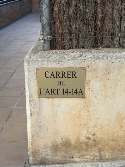 Street sign "Carrer de L'art 14-14A" attached to a weathered concrete wall in Rubí, Spain. A woven reed fence is visible above the wall. Tiled pavement is below. The image is oriented with the text running vertically.