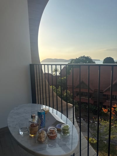 An overhead view from a balcony in Cenang Beach, Malaysia, captures a late afternoon or early morning scene. In the foreground, an oval white marble table holds an empty clear glass, a small bottle of Monkey Shoulder blended malt scotch whisky, a blue and white "Revive" branded soda can, an open white book or menu, and three small clear glass jars containing snacks (light-colored, reddish-brown, and green items). The balcony floor is light-colored decking. A black metal railing with vertical bars spans the midground, beyond which traditional buildings with reddish-brown tiled roofs and lush green tropical foliage are visible. In the background, a body of water with distant islands meets a sky displaying a yellow-orange to light blue gradient, indicative of low sun angle. No individuals are present.