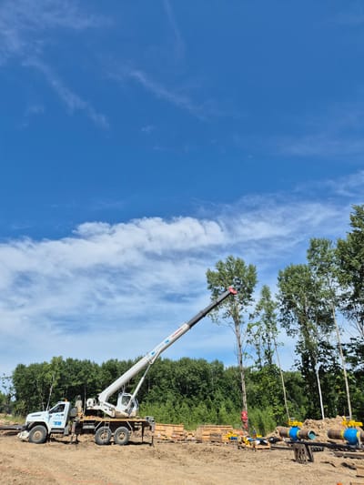 In Taezhnoe, Russia, on a clear day with a blue sky and scattered clouds, a construction scene unfolds. A white crane truck is positioned on uneven, sandy terrain, its boom extended upwards and diagonally to the right. The truck has a white cab with blue decals and appears to be a utility vehicle. Various construction materials, including wooden crates and large blue and yellow valves connected to pipes, are scattered around the site. A dense forest of green trees forms the backdrop, with some taller trees standing out in the foreground. The ground is a mix of dirt and sand, with some orange marking lines visible. The overall impression is a construction project in progress in a rural, wooded area.