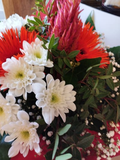 A close-up, vertical shot captures a vibrant bouquet of flowers. The arrangement features a prominent red, spiky flower at the top, flanked by fiery orange gerbera daisies. Interspersed throughout are delicate white chrysanthemums with pale green centers. The bouquet is complemented by lush green foliage and small white filler flowers, possibly baby's breath, adding a touch of airy texture. Some of the flowers are wrapped in red paper. The overall impression is one of a celebratory or decorative arrangement, likely indoors given the soft lighting. The image evokes a feeling of freshness and natural beauty. No text is visible. The location context indicates this photo was taken in Zikhron Yaakov, Israel.