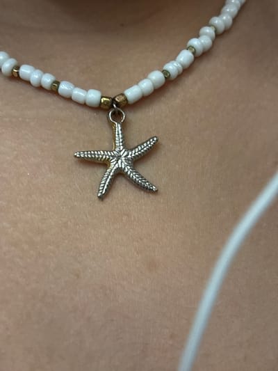 A close-up shot captures a silver starfish pendant hanging from a white beaded necklace. The necklace is composed of small, round white beads interspersed with tiny gold beads. The pendant is detailed, with raised lines creating the texture of a starfish. The necklace rests on a person's skin, which appears to be a tan complexion. The background is out of focus, showing a hint of a white strap or fabric on the right side of the image. The location of this image in Bistrice, Montenegro, cannot be determined from the visual information provided.