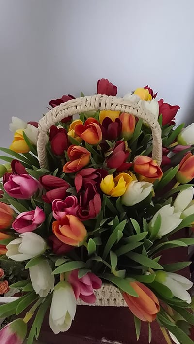 The image features a close-up, vertically oriented shot of a basket overflowing with artificial tulips. The tulips display a vibrant and diverse color palette, including shades of red, yellow, orange, pink, and white. Green foliage is interspersed throughout the arrangement, adding to the fullness and naturalistic appearance of the bouquet. The basket itself is made of a woven, light-colored material, with a thick rope-like handle. The background is divided into two sections: a plain, light gray surface on the left and a darker, reddish-brown surface on the right, possibly indicating furniture or a wall. The lighting appears to be artificial or from an overcast sky, creating a soft illumination across the subjects. No people are visible, and the scene is static, focusing solely on the floral arrangement within the basket. The image does not provide any specific cues about the time of day, weather, or location beyond what might be inferred from the artificial nature of the flowers. There is no visible text in the image.