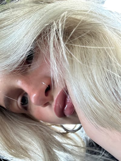A close-up, high-angle portrait displays an individual's head tilted, with the face largely framed by platinum blonde hair. The subject gazes directly at the camera. A small silver hoop nose ring is visible in the left nostril. The lower lip is full, glossy, and appears slightly parted. The left eye is partially obscured by hair, revealing a dark pupil, while the right eye shows a dark iris and pupil. Fair skin tone with slight redness around the nose is present. A metallic, circular earring is partially visible on the lower right side of the frame. The background is out of focus and undifferentiated, providing no discernible environmental details. The exact location within Saanich, Canada, cannot be determined from the image.