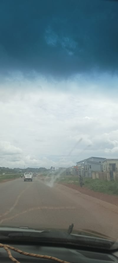 The image shows a road view in Ozalla, Nigeria, from inside a vehicle. The windshield is partially obstructing the view. A road stretches into the distance with a white pickup truck driving on it. Buildings line the right side of the road. A person walks along the roadside. The sky is overcast.