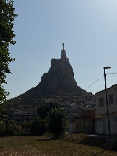 The image shows a daytime, outdoor scene in Monteagudo, Spain, dominated by a prominent rock formation topped with a statue of Christ the King. The statue is positioned high on the rocky peak, with its arms outstretched. The rock formation itself is steep and rugged, with some visible remnants of fortifications or structures on its upper slopes. Below the rock formation, a cluster of houses and buildings forms the town of Monteagudo, nestled on the hillside. The foreground features a grassy area with dry grass and some trees on the left side. A street lamp and parts of buildings are visible on the right side of the image, including a white building with windows and a ribbed garage door. The sky is clear and blue.