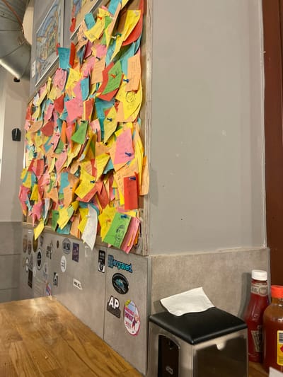 The image, rotated 90 degrees clockwise, shows a vibrant wall densely covered with numerous colorful sticky notes, primarily yellow, pink, blue, and red. These notes appear to be pinned to the surface, many displaying handwritten messages, drawings, or symbols. Above this wall, to the left in the current orientation, is a plain gray wall or counter surface.
To the right side of the image, there is a gray countertop with two condiment bottles: a red ketchup bottle and an orange-brown bottle, possibly hot sauce or another dressing. Next to them is a black napkin dispenser with white napkins. Below this countertop, a vertical panel is adorned with an assortment of stickers. These stickers feature various designs, logos, and some legible text, including "engraved.", "AP FILMS", "VODKA WORRY" (on an oval yellow sticker with a cartoon character), and "WOZ SE." (or "WOZ SF.").
On some of the colorful notes, partially readable text includes phrases like "... I love you...", "...BVB...", "...PULP, AXEL, DOR, OCTOBER...", and "Min Bar".
Further to the bottom left, a section of a framed artwork or map is partially visible, along with some metallic ductwork. The lower right portion of the image reveals a warm-toned wooden floor. The overall scene suggests a lively interior environment, likely a cafe, restaurant, or bar, where patrons are encouraged to leave their mark.
