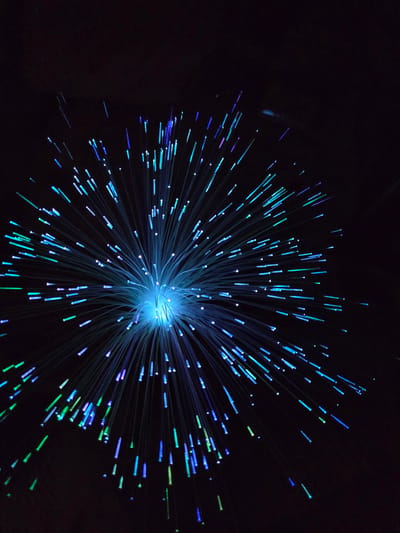 The image depicts a fiber optic light display against a dark background. The light source is centrally located and emits numerous strands of light, predominantly blue, with some strands exhibiting green and purple hues. The light strands radiate outwards from the central source, creating a starburst effect. The background is a dark void, allowing the illuminated strands to stand out prominently. There are no people or other objects visible in the scene. The location of the image is unknown.