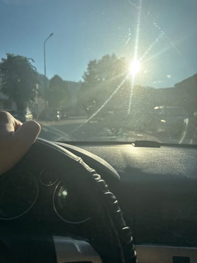 The image shows a first-person perspective from inside a vehicle, with a hand on the steering wheel. The sun is bright and high in the sky, causing glare and lens flare on the windshield. The view through the windshield shows a street scene with trees and buildings, suggesting an urban or suburban environment. There are faint water spots or smudges on the windshield. The dashboard of the car is visible in the foreground, including parts of the instrument cluster. The time of day appears to be daytime, likely late morning or early afternoon, given the sun's position. The location is specified as Smilyan, Bulgaria.