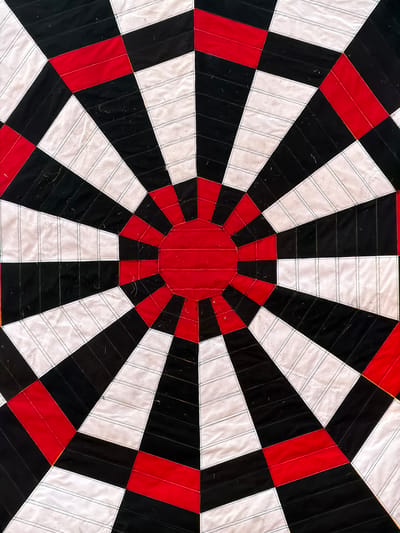 The image is a close-up, top-down view of a quilted fabric object with a circular, radial pattern. The pattern is composed of alternating wedges of black and white fabric radiating outwards from a central red circular area. Interspersed between some of these wedges are rectangular sections of red fabric. The white fabric sections have visible stitching lines running horizontally across them, creating a striped effect. The object appears to be laid flat, possibly on a floor or table, and is part of a larger structure or design. There are no people, animals, or other environmental elements visible in the frame. The lighting is even, suggesting an indoor setting or diffused daylight. The context of Ракітніцкі сельскі Савет, Belarus is not determinable from the visual information provided in the image.