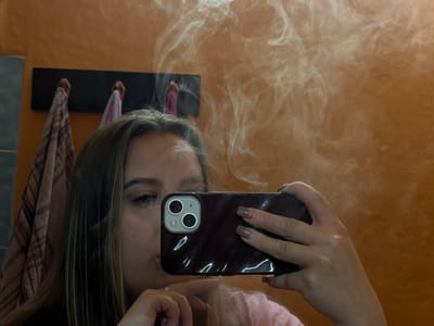 A person is taking a selfie in a mirror, partially obscuring their face with a dark-colored smartphone. Smoke or vapor is prominent in the reflection. The background includes a textured orange wall, a dark-colored rack with hanging towels, and a glimpse of a blue-tiled area. The location is not mentioned in the image.