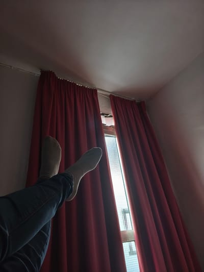 The image presents an indoor scene captured from a low-angle perspective, looking upwards towards a window and ceiling. The central subjects are a person's legs, dressed in dark blue jeans and light-colored, possibly off-white, socks, resting in an elevated position, suggesting relaxation on a bed or couch.

Behind the person's feet, a window is partially covered by two rich, dark red curtains. The curtains are mostly drawn, but a vertical gap in the center reveals a brightly lit exterior, possibly through a frosted or patterned glass windowpane. The strong light from outside indicates it is daytime. The interior of the room appears dimly lit, with soft shadows on the pale ceiling and walls, creating a cozy and tranquil atmosphere. Above the curtains, a white curtain rod is visible.

No specific activities are depicted other than the person relaxing with their feet up. There is no visible text within the image. While the image itself does not contain explicit location cues, the provided context indicates the scene is located in Moaña, Spain. The overall impression is one of quiet repose and comfort.