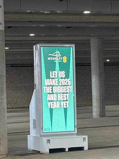 An indoor scene displays a brightly lit digital advertisement board mounted vertically on a structural element, possibly a wall or pillar, within a public concourse or building. The background shows neutral-colored architectural features, including horizontal beams and textured walls, typical of a large venue. No people are visible.
The screen exhibits a teal green background with white text and logos. At the top, the "Wembley" logo is prominently displayed, followed by "CONNECTED BY EE," with the EE logo also visible. The main message on the board reads: "LET US MAKE 2025 THE BIGGEST AND BEST YEAR YET." The text is large and sans-serif, arranged vertically, indicating an aspirational message for the upcoming year. Below the text, two green triangular shapes point upwards. In the bottom right corner of the screen, faint small icons are visible, likely generic display controls or status indicators. The environment appears well-lit by artificial lighting. Given the "Wembley" branding, the location is confirmed to be at Wembley in the United Kingdom, likely within Wembley Stadium or an associated complex.