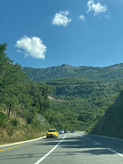 A bright, sunny day is captured from a moving vehicle on a road that winds through a lush, mountainous landscape. The road is paved asphalt with distinct white and yellow lane markings. In the foreground, a bright yellow car is driving in the right lane, followed by several other vehicles. To the left of the road, there is a steep, vegetated embankment with scattered trees and bushes, and a roadside sign. To the right, the road curves around a rocky, tree-covered mountainside. In the background, dense green forests cover the slopes of imposing, rugged mountains under a clear blue sky dotted with a few fluffy white clouds. The location appears to be in the mountainous region surrounding Markovići, Montenegro, characterized by its steep terrain and vibrant vegetation.