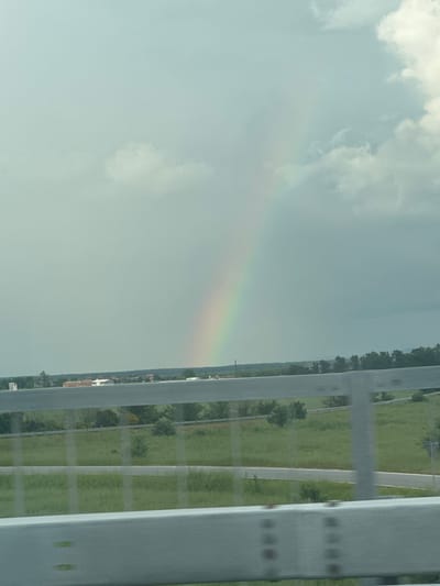 A faint rainbow is visible in the sky. A highway runs parallel to the sky, separated by a guardrail. There is green vegetation beyond the highway. The image's location is unknown.