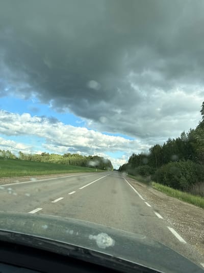 The image depicts a view from inside a moving vehicle traveling on a rural road near Beržiniai, Lithuania. The road is asphalt, marked with broken white lines indicating lanes. The weather appears unsettled, with a mix of dark storm clouds and patches of blue sky. Vegetation, possibly trees and fields, lines both sides of the road. Droplets on the vehicle's windshield suggest it is raining or has recently rained. There are no visible people or other vehicles in the immediate vicinity.