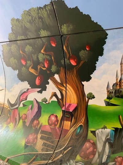 This media file displays a close-up, oblique view of a colorful, fantasy-themed mural or artwork. The artwork appears to be composed of multiple square panels, with visible seams between them. The style is illustrative and cartoonish, featuring a whimsical landscape with a large, stylized tree as a central element. The tree has a thick, gnarled trunk with warm, brown and orange tones, and its branches are adorned with bright red, spherical fruits or objects.
The backdrop includes a bright blue sky with wispy white clouds. To the upper right, a whimsical castle with pointed spires is depicted against a vibrant green, rolling landscape. Scattered throughout the scene are various fantastical elements, including mushroom-like objects with reddish-brown caps dotted with darker circles, and abstract, geometric shapes in shades of pink and purple. One prominent detail is a small, cube-shaped structure, possibly a house or a building, nestled within the tree trunk. The artwork has a bright, cheerful, and imaginative feel, evoking a fairytale or fantasy setting. There is no visible text within the image. The lighting suggests it could be daytime, with clear weather. The location context provided is Odivelas, Portugal, suggesting this artwork is located in that area.