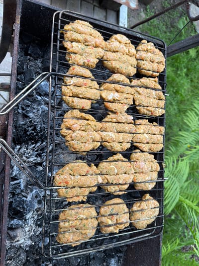 In an outdoor setting, likely a backyard in Каменка, Russia, a metal grill rack is positioned over glowing charcoal. The grill is laden with numerous raw, oval-shaped patties, appearing to be made of ground meat or a similar mixture, with visible flecks of herbs and red peppers. These patties are arranged in several rows on the wire rack. To the left of the grill, the dark, charred remains of burnt wood are visible. The background features blurred green foliage, suggesting a natural or garden environment. No people are present in the image.