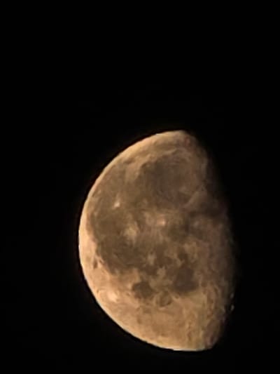 A close-up, dimly lit, upward-angled shot captures a portion of the moon against a black sky. The moon's surface displays variations in color, ranging from light brown to dark tan, with visible craters and texture. The right side of the moon is illuminated, while the left side is in shadow. There are no people, animals, buildings, or any other discernible objects or elements in the image besides the moon and the night sky. The image does not provide any information about the city of Икское Устье, Russia, nor its specific location within it.