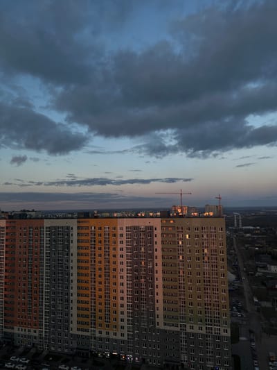 Captured from an elevated perspective in Анкудиновка, Russia, the image depicts a high-rise apartment building juxtaposed against a dramatic sky. The building, painted in horizontal bands of green, gold, gray, and brown, dominates the right side of the frame. Numerous windows punctuate its facade, with some emitting light, suggesting occupancy. To the left, a heavily clouded sky casts an ominous tone. Two red construction cranes are visible in the mid-ground. Distant buildings and infrastructure hint at the city's expanse. The composition is oriented with the sky and horizon running vertically.