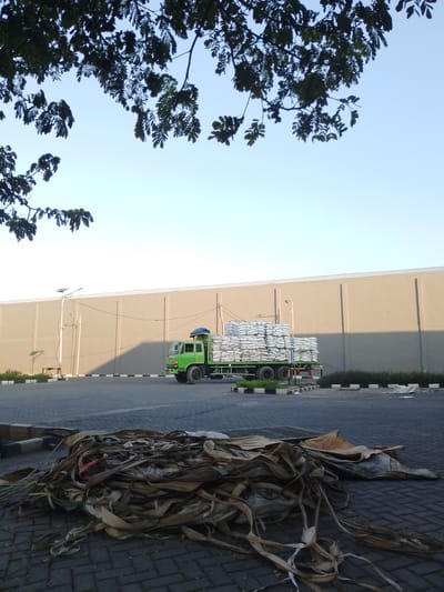 In Banyuwangi, Indonesia, a bright green truck is parked on a paved lot in front of a large, light brown industrial building. The truck is loaded with many sacks, likely containing goods, all bundled together and covered with a blue tarp. To the left of the truck, a lamppost with solar panels stands tall. In the foreground, a pile of discarded brown and tan woven material, possibly sacks or tarps, is scattered across the pavement. Overhead, the dark green leaves of a tree branch partially obscure the clear blue sky. The scene appears to be an industrial or storage area, likely during daylight hours.