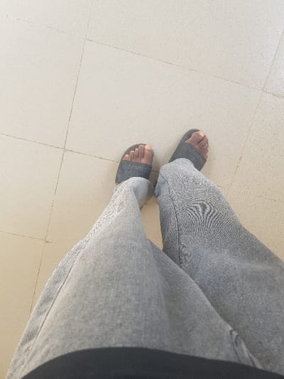 The image depicts a top-down view of an individual's lower torso, legs, and feet. The specific location within Panhauya, Nigeria, is not discernible from the visual evidence. The subject is wearing light-grey, loose-fitting denim trousers and dark grey or black slide sandals with textured straps on their feet. The skin on the visible feet is dark-toned. A dark-colored waistband is visible at the bottom edge of the frame. The individual is standing on a light beige or off-white tiled floor consisting of square tiles with thin, darker grout lines forming a grid pattern. The floor appears clean and uniformly laid. No other individuals or interactive elements are present in the scene.