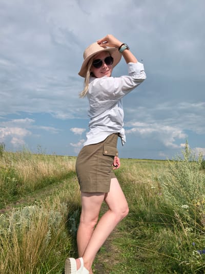 A woman wearing a white button-down shirt, khaki shorts, a straw hat, and sunglasses stands in a grassy field under a partly cloudy sky. She has her left hand on her hat and her right arm is bent at the elbow. She is looking towards the camera with a slight smile. The field appears to be a rural area with tall, dry grass and scattered green bushes. The sky is a mix of blue with white and grey clouds. The location is identified as Kuzminovka, Russia.