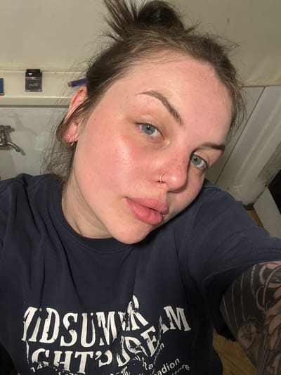 A woman with fair skin, light blue eyes, and brown hair pulled back in a bun is shown in a close-up, selfie-style photograph. She has a nose ring in her left nostril and is wearing a dark navy-blue T-shirt with white lettering. A portion of a tattoo is visible on her right arm. The background appears to be a tiled bathroom, with a sink and wall accessories visible. The location in the city of Chornomorskoe, Ukraine cannot be independently confirmed from the provided image.