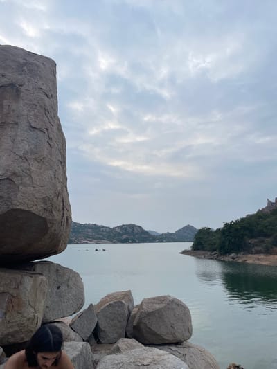The image displays a rugged natural landscape near a body of water in Sanapur, India, under an overcast sky. In the immediate foreground, a massive, weathered granite boulder dominates the left side of the frame, stacked upon other large boulders. Partially visible at the bottom-left, an individual with dark, shoulder-length hair is seated or crouched amongst these rocks, their head angled downwards.

A calm body of water, likely a lake or reservoir, occupies the midground, its surface showing slight ripples and reflecting the muted light of the sky. The far bank of the water reveals a series of rolling hills and mountains, characterized by numerous exposed granite rock formations and sparse green vegetation. On the right side of the water, the shoreline transitions from rocks to a denser collection of green trees and bushes, extending into similar rocky hills in the distance. The sky is uniformly covered with light grey clouds, indicating diffuse lighting conditions.