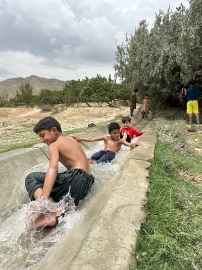 In Kan Mehtarzai, Pakistan, children are enjoying themselves on a makeshift water slide. A concrete channel, serving as the slide, is filled with flowing water, creating splashes as the children go down. The first boy, bare-chested and wearing dark pants, is in the foreground, his face lit with a wide smile as water sprays around him. Behind him, a boy in a red shirt and dark pants slides with his arms outstretched, also smiling. Another boy, further up the slide and out of focus, is also visible. Several other individuals are present in the background, including adults standing and observing. The environment appears to be rural, with dry, dusty terrain, scattered trees, and distant hills under a cloudy sky. The scene captures a moment of playful enjoyment amidst a simple, natural setting.