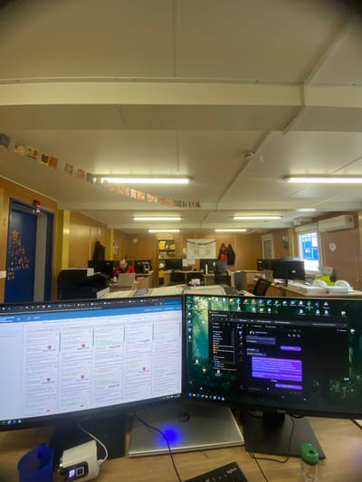 The image shows an office environment with multiple computer monitors displaying various programs and applications. The perspective is slightly tilted and the scene extends into the background, showing more desks and office equipment.

The foreground is dominated by computer monitors, a laptop, and office supplies such as a mouse and a small electronic device showing a digital reading of "70." The screens display what seems to be work-related applications. One shows a messaging application with purple text bubbles, and the other displays tables filled with data and text in Russian.

In the background, there's a glimpse into other parts of the office. People are working at desks, and there's a banner in the center of the image. Decorations hang from the ceiling. 
There is no definitive information to confirm the location in the city of Luzhitsy, Russia.