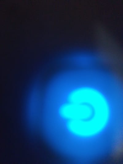 The image is a close-up, out-of-focus shot of a bright blue circular light against a dark background. The light appears to be the power indicator of an electronic device. There are no people or discernible objects other than the light source itself. The location in Rumuomasi, Nigeria, is not visually represented in the image.
