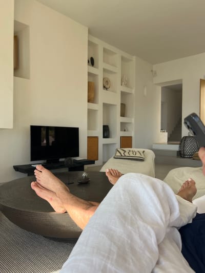 The image displays an interior view of a modern living space, specifically identified as being in Agios Ioannis Diakoftis, Greece. The scene is observed from a low angle, focusing on the immediate foreground occupied by the bare legs and feet of two individuals. The individual on the left has crossed, tanned legs. The individual on the right, wearing white linen trousers, has uncrossed, tanned feet; their hand, holding a dark smartphone, is visible in the upper right corner, indicating the camera operator.
A large, circular, dark-textured coffee table is centrally positioned, bearing a small dark sculptural object and a clear glass decorative item. Beyond the coffee table, a black flat-screen television rests on a low, dark media console. The prominent white wall behind the television features multiple built-in recessed shelves, filled with various decorative objects including vases, discs, and abstract sculptures in dark and light tones. To the right of the television setup, a light-colored chaise lounge with a black and white patterned pillow is visible. In the background, an archway leads to an area with a visible staircase and a large, dark, intricately designed lantern. The room is brightly illuminated by natural light, emphasizing its minimalist aesthetic and elements consistent with Cycladic architectural style.