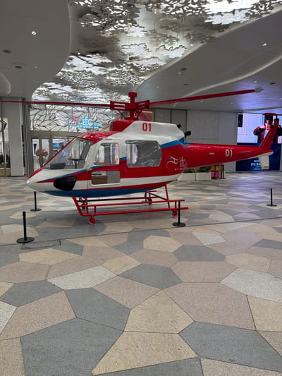 The image depicts a static, red and white helicopter model situated inside a building, likely a shopping mall, characterized by modern architecture and design. The helicopter, bearing the number "01" on its tail and "10" on its side, is elevated on a red support frame. The environment features a tiled floor with a patchwork pattern of neutral tones. In the background, there is a wall decorated with white, floral-shaped elements. The IFC mall can be seen through the glass doors. Based on the information inferred from the image, the location is likely in the Jianye District, China, specifically near the IFC mall.