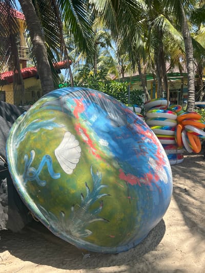 This image captures an outdoor scene on a sunny day, likely in Hoi An, Vietnam, given the presence of palm trees and what appears to be a tropical setting. In the foreground, a large, colorful, spherical object dominates the frame. This object is painted with a mural depicting marine life and underwater scenes, including a white seashell and blue coral-like shapes against a background of green and blue, with streaks of red paint. The object is resting on sandy ground, partially covered by a black tarp on its left side. Behind this prominent object, a stack of inflatable rings, in various colors like orange, yellow, blue, and white, are piled high. The rings are neatly arranged and suggest a place associated with recreation or a beachside establishment. In the background, several palm trees with their fronds are visible, some casting shadows on the sand. Further back, buildings with red-tiled roofs and yellow walls can be seen, hinting at structures possibly associated with resorts or shops. The overall atmosphere is relaxed and suggestive of a tourist destination. There are no people visible in the image.