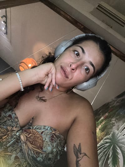 The image shows a woman lying down, possibly on a bed or couch, wearing headphones. She is looking upwards and to the side with a thoughtful or contemplative expression. Her right hand is resting on her chin, with her fingers splayed. She is wearing a patterned top and a necklace with a script name on it. A tattoo of a hummingbird is visible on her upper arm. The background features a wall with a tropical leaf pattern and a lamp. The lighting suggests it might be evening or indoors. The location context is Khan Daun Penh, Cambodia.