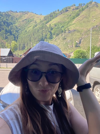 A woman is taking a selfie outdoors in Manzherok, Russia. She is wearing a light-colored bucket hat, dark-rimmed glasses, and a striped shirt. Her left arm is raised, and she appears to be looking at the camera with a playful expression. In the background, a mountainous landscape covered in trees and vegetation is visible. There are also some buildings, a street, and a light pole. The scene is illuminated by bright sunlight.