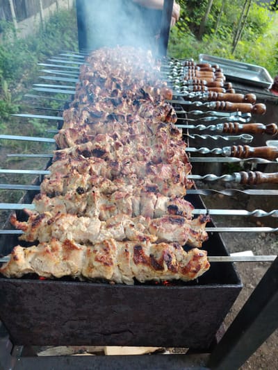This image captures a close-up view of numerous skewers of meat being grilled over an open fire in a rustic, outdoor setting. The meat, appearing to be pork, is cut into bite-sized pieces and threaded onto long metal skewers. Some pieces are lightly browned, while others are developing a more caramelized crust, indicating they are in various stages of cooking. Wisps of smoke rise from the grill, obscuring the background slightly.

The grill itself is a dark, weathered metal brazier, suggesting it has been used many times. Several other skewers with wooden handles are visible to the right, resting on the edge of the grill, some appearing to be prepared with meat and others empty. In the background, lush green foliage and trees can be seen, with hints of a wooden fence on the far left, suggesting a rural or suburban location. The lighting suggests it could be daytime, possibly late afternoon, given the soft natural light.

The scene evokes a sense of outdoor cooking and perhaps a gathering or event. There is no visible text in the image. The focus is entirely on the appetizing grilled meat and the process of cooking it.