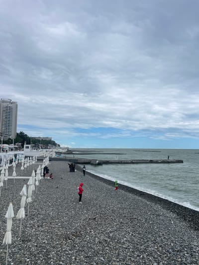 Captured on a beach in Bolshoy Sochi, Russia, the image presents a pebble-strewn coastline meeting the sea under a sky of scattered clouds. A concrete pier stretches into the water. Several people are positioned along the beach, including one figure on the pier. In the foreground, white umbrellas line the shoreline. A multi-story building stands in the distance behind the beach umbrellas.