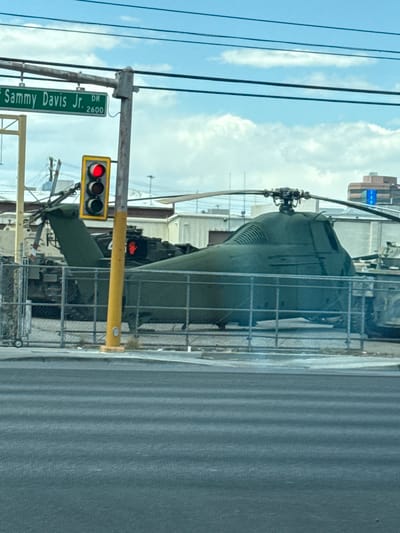 The image shows a military helicopter on display near a street corner in Las Vegas, Nevada, USA. The helicopter is green and partially obscured by a metal fence. A street sign reads "Sammy Davis Jr. Dr. 2600" indicating the location. A traffic light is visible, showing a red signal. The sky is cloudy.