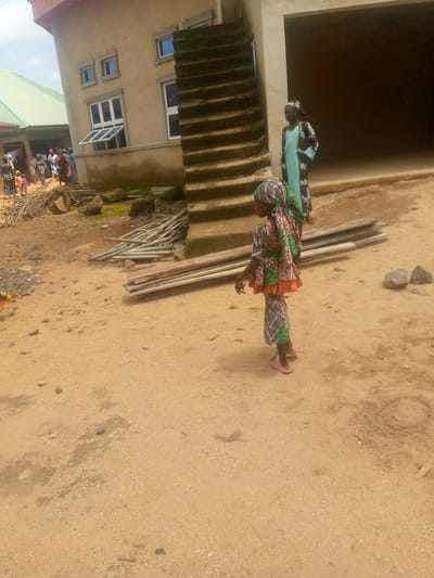 In Madakiya, Nigeria, a young girl wearing a brightly patterned dress and headscarf stands on sandy ground. She is facing left, with her left arm extended. Behind her, a woman in a patterned blue and brown dress and headscarf stands near a large building with an open entrance. To the left of the building, several people are gathered, and some construction materials, including metal pipes and wooden planks, are visible on the ground. The environment appears to be an outdoor area near a building under construction.