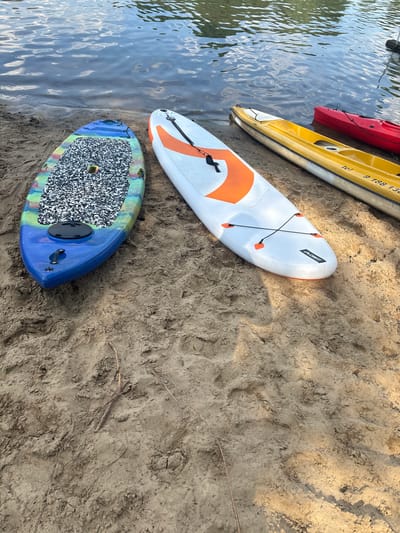 In Kobyla Góra, Poland, on a sandy shore next to a body of water, there are several watercraft lined up. A blue paddleboard with a colorful, speckled design rests on the sand, its nose pointing towards the water. To its right, a white paddleboard with an orange graphic pattern is also positioned on the sand, parallel to the blue one. Further to the right, a yellow kayak is visible, followed by a red kayak. The water exhibits ripples and reflections of the sky. The sand is coarse and shows signs of being disturbed, with small sticks and debris scattered across its surface. The scene depicts a tranquil setting by the water, with recreational equipment ready for use.