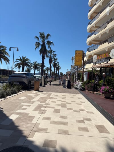 The image depicts a street scene in Fréjus, France. A row of tall palm trees lines the left side of the image, reaching towards a clear blue sky. A multi-story building with a facade of protruding balconies stands adjacent to the trees. A yellow sign for "RESTAURANT ASIA" is visible, indicating an Asian cuisine establishment. 

On the right side of the image, a patterned sidewalk is evident, featuring light and dark rectangular tiles in a seemingly random arrangement. The perspective suggests the photo was taken at street level. People can be seen walking along the sidewalk in the middle distance. A parked car is visible at the curb. The atmosphere suggests a sunny day in a coastal city.