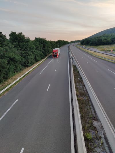This is a high-angle shot of a highway in Marchaux-Chaudefontaine, France, during dusk. A red semi-truck is traveling on the right lane of the highway, heading away from the camera. The highway is a multi-lane road with concrete barriers separating the opposing directions of traffic. Green trees line the left side of the highway, and a forested hill is visible on the right. The sky is a soft gradient of pink and orange, indicative of sunset. The highway markings are visible, including white dashed lines separating lanes and solid white lines along the shoulders. There are other vehicles in the distance on the highway, but they are too small to be identifiable.