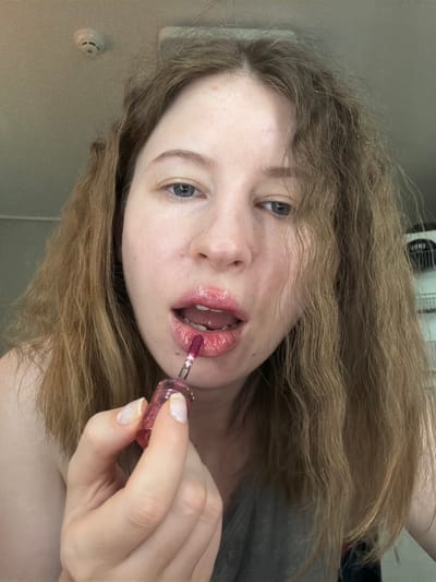 A woman with light brown hair is applying dark pink lip gloss to her lips. She is holding the lip gloss wand in her right hand and appears to be looking into a mirror. The background is out of focus but appears to be an indoor setting with a smoke detector on the ceiling. The image is cropped to focus on the woman's face and hand. This image was captured in Dongducheon-si, South Korea.