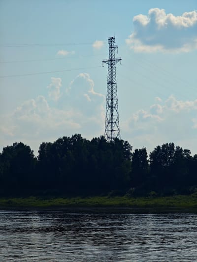 An outdoor, low-angle, landscape shot captures a power line tower silhouetted against a bright, hazy blue sky with scattered white clouds. The tower is a tall, lattice-metal structure, with three sets of horizontal power lines stretching from its upper section. Below the tower, a dark, dense line of trees forms a solid band across the middle of the frame. In the foreground, a wide, dark river occupies the bottom half of the image, its surface rippled by the wind, reflecting the muted light of the sky. The scene appears to be set in a natural, undeveloped area, possibly on the outskirts of a settlement like Verkhnepashino, Russia, given the presence of power infrastructure in a predominantly natural setting. There are no people or animals visible in the frame.